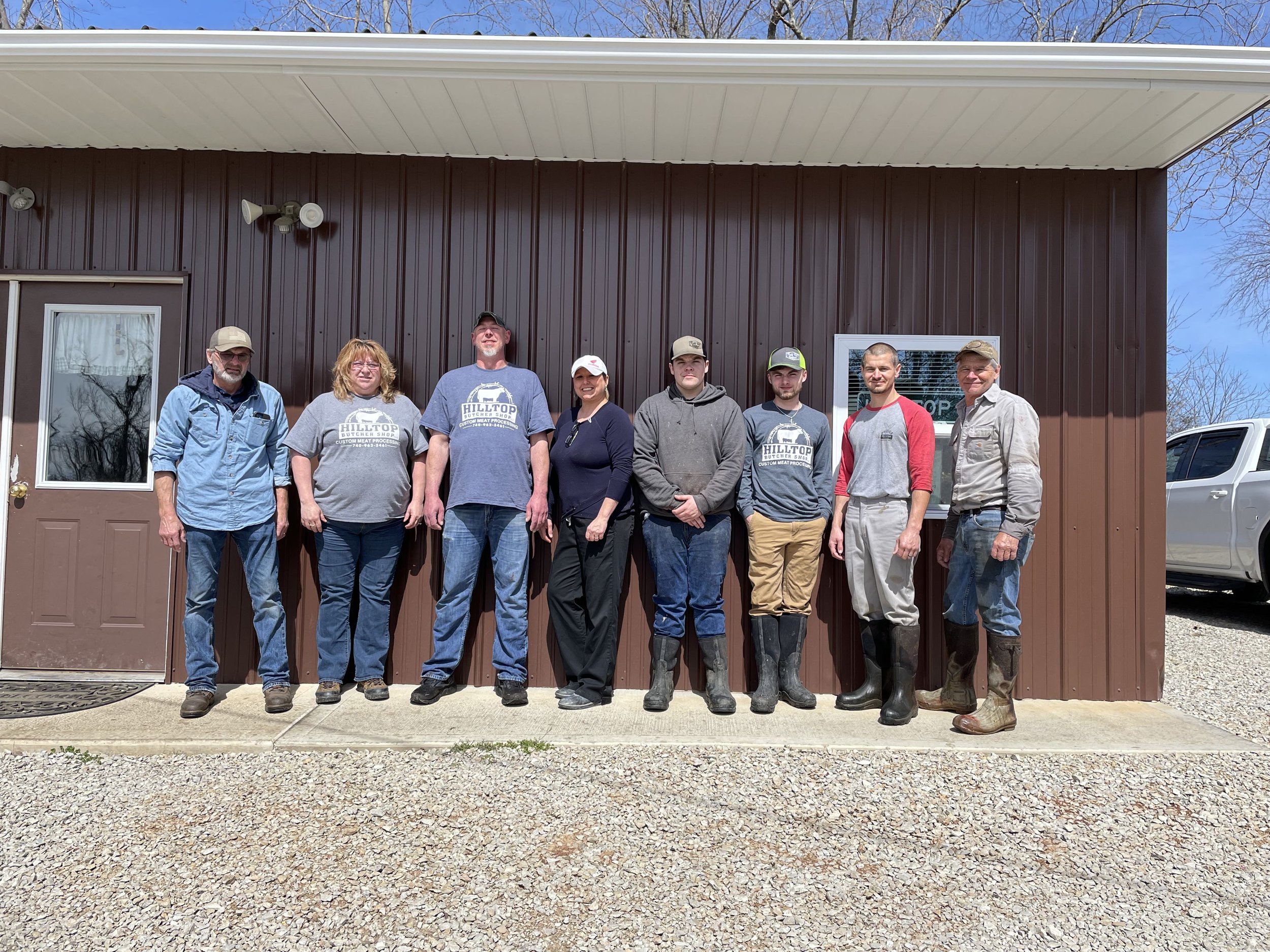 Hilltop_Butcher_-_IMG_1130 Group of nine people standing in front of a brown metal building on a sunny day, wearing casual clothing and boots, representing a community or agricultural organization.